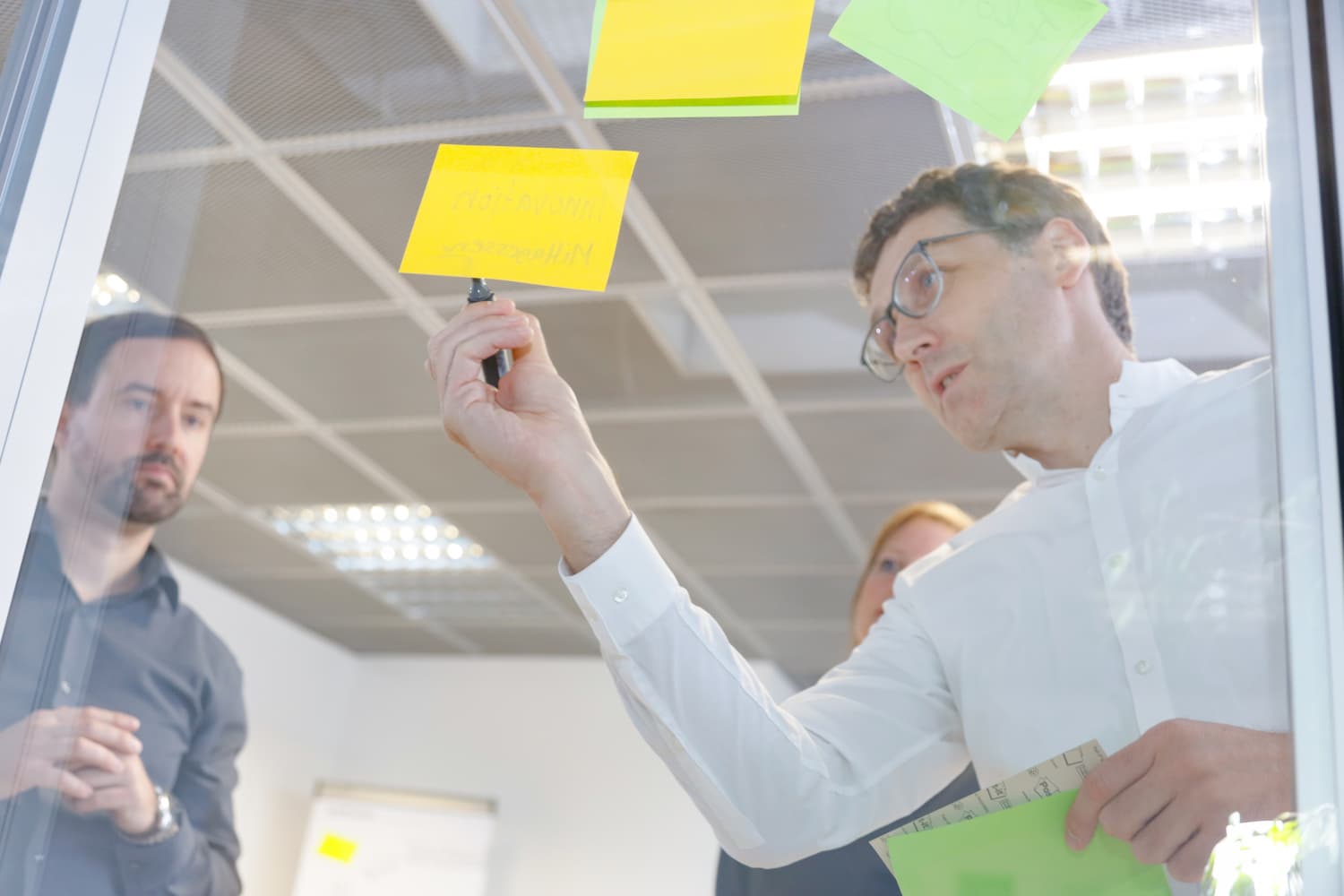 Bild-Text-Streifen Coach schreibt etwas auf ein gelbes Post-it an einer Glaswand